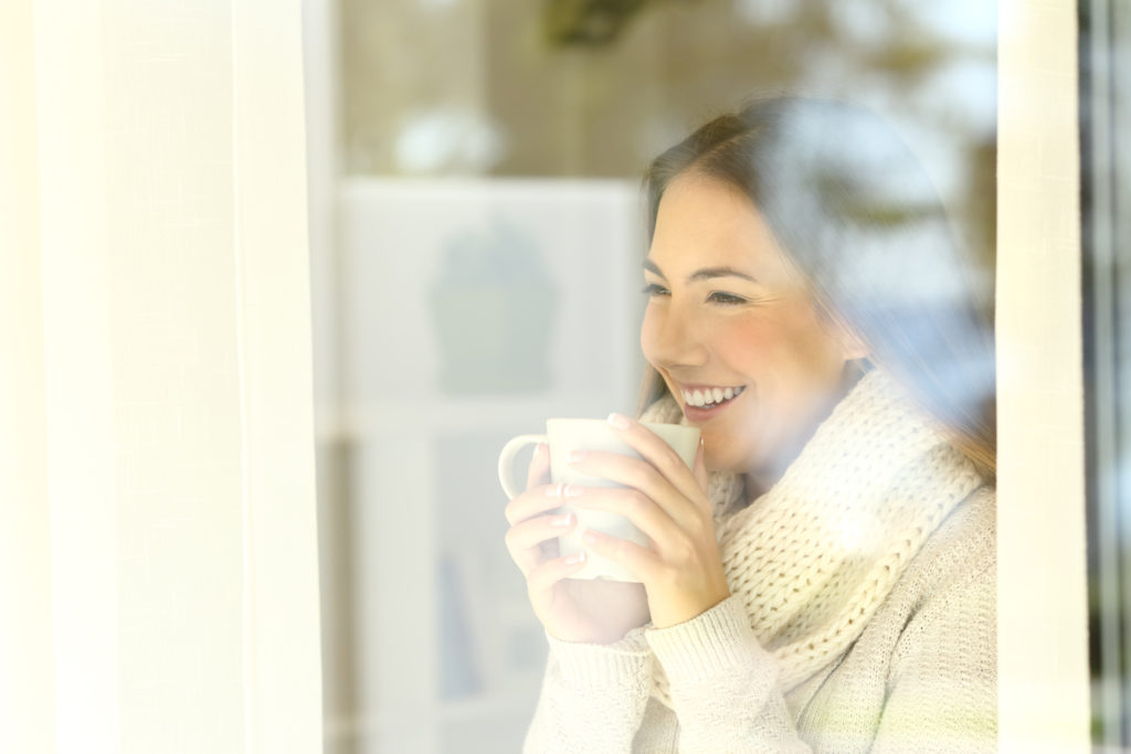 Happy lady looking through a window holding a coffee mug standing at home in winter