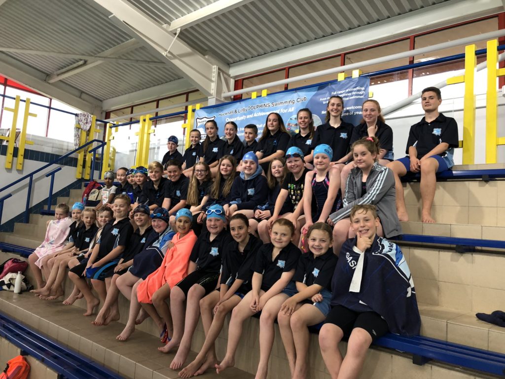 Young swimmers from the Hucknall and Linby Dolphins sit in tired seating by the pool to have a club photo.